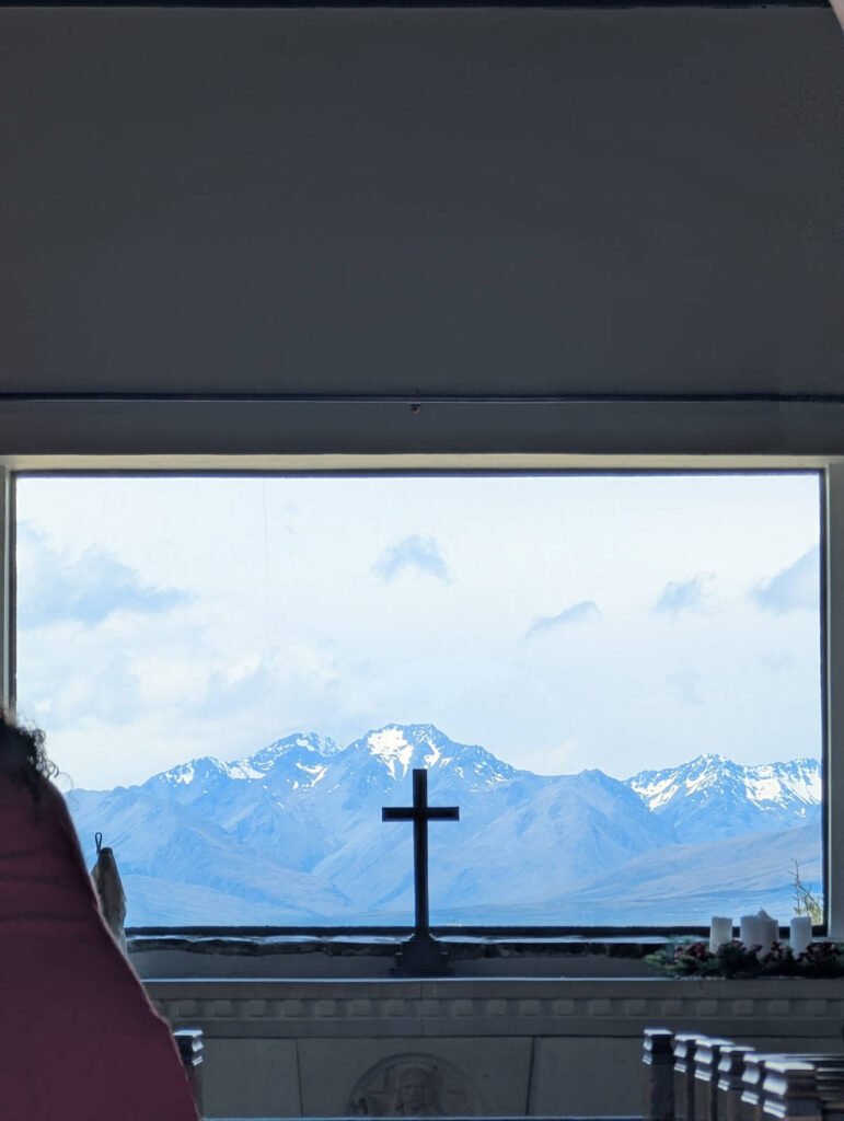 View from inside the Church of the Good Shepherd, where a simple cross on the altar is silhouetted against a large window framing snow-capped mountains.