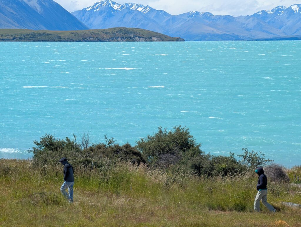 Two people walk along the windswept shore of Lake Tekapo, its distinctive turquoise glacial waters choppy beneath snow-capped Southern Alps mountains.