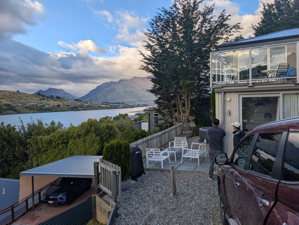 A hillside Airbnb in Queenstown with a gravel patio and white outdoor furniture, overlooking Lake Wakatipu and surrounding mountains under a dramatic evening sky.