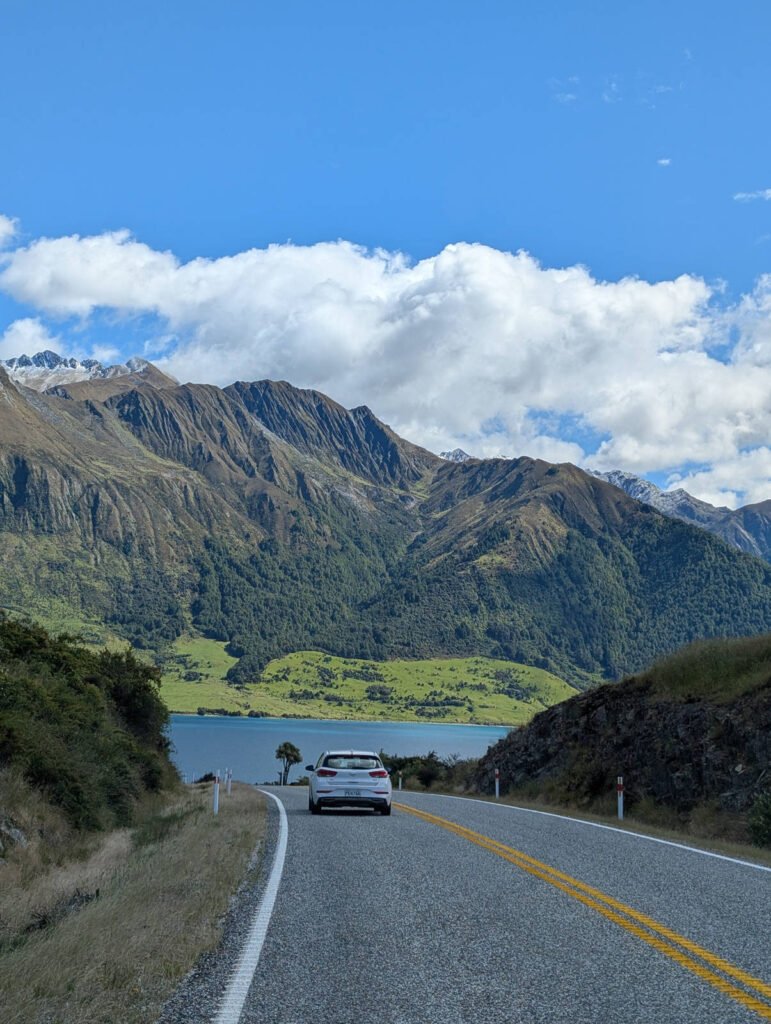 A white car travels along a winding road beside a vivid blue lake, with bright green pastures and towering mountains — some snow-capped — under a sunny sky in New Zealand's South Island.
