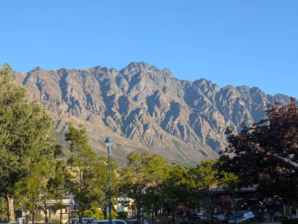 The jagged peaks of The Remarkables mountain range tower over Queenstown's town centre, framed by leafy trees under a clear blue sky.